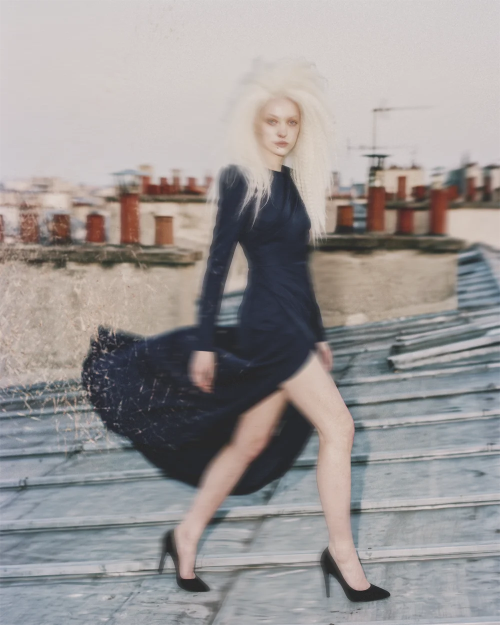 Dreamy soft-focus editorial of a blonde model in a navy dress walking across a rooftop among chimneys – Sbranzo Alessandro Valori Studio Vago
