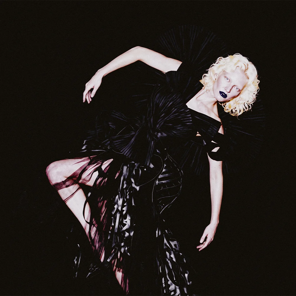 Editorial of Elena arched in a radial pleated black gown with raised arm and blue lips against a black background – Sbranzo Alessandro Valori Studio Vago Elena