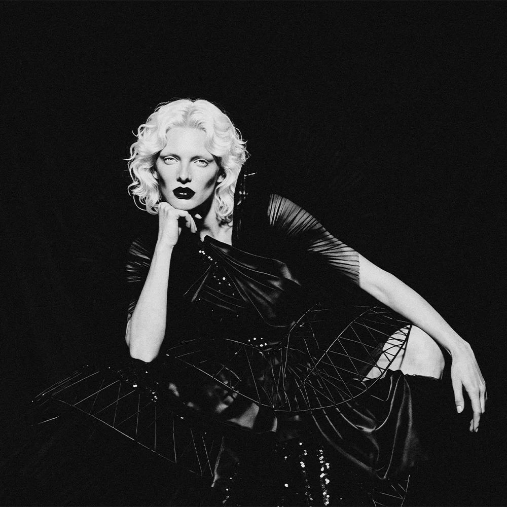 Black and white editorial portrait of Elena seated with hand under chin in a sculptural black corset gown – Sbranzo Alessandro Valori Studio Vago Elena