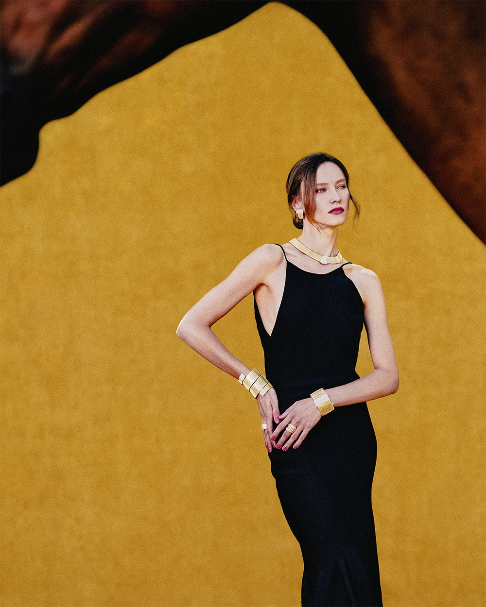 Fashion jewelry editorial 999 – model in a black dress wearing a gold rigid necklace, wide bracelets and ring against a yellow textured horse backdrop – Sbranzo Alessandro Valori Studio Vago Valeria Miro