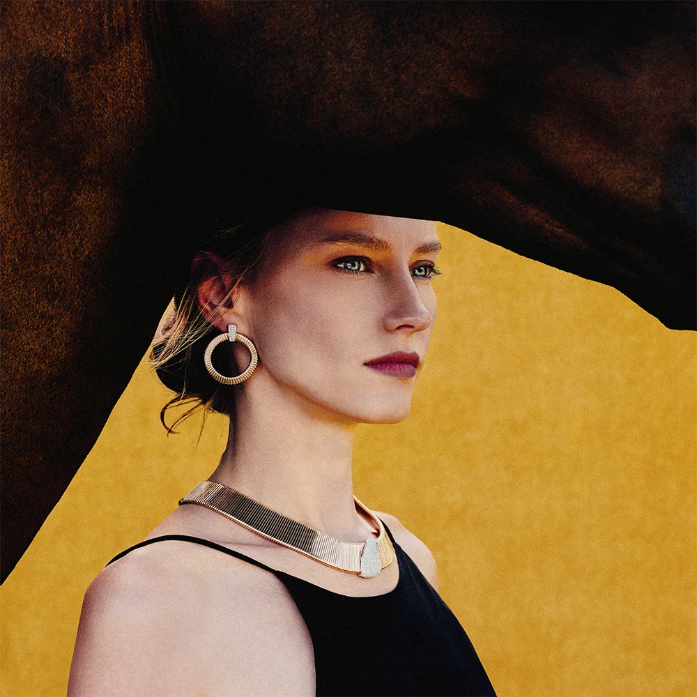 Fashion jewelry editorial 999 – close-up portrait of a model wearing a gold hoop earring and rigid necklace against a yellow horse backdrop – Sbranzo Alessandro Valori Studio Vago Valeria Miro