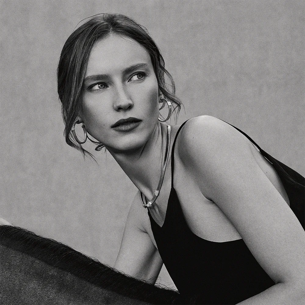 Black and white fashion jewelry editorial 999 – portrait of a model with windswept hair wearing hoop earrings and a necklace next to a horse saddle – Sbranzo Alessandro Valori Studio Vago Valeria Miro