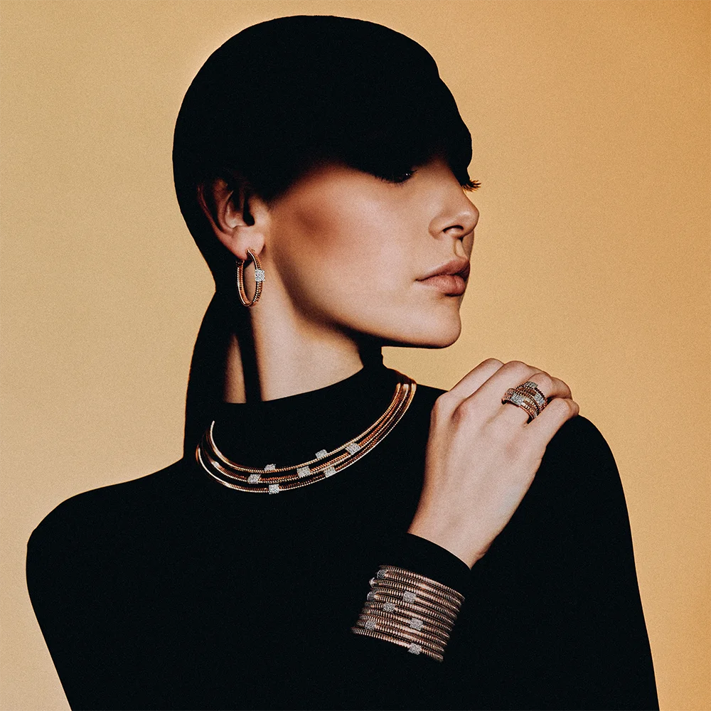 Fashion jewelry editorial 999 – model in profile wearing a hoop earring, multi-strand necklace, wide stacked bracelet and ring against a camel background – Sbranzo Alessandro Valori Studio Vago Viola Sgriscia
