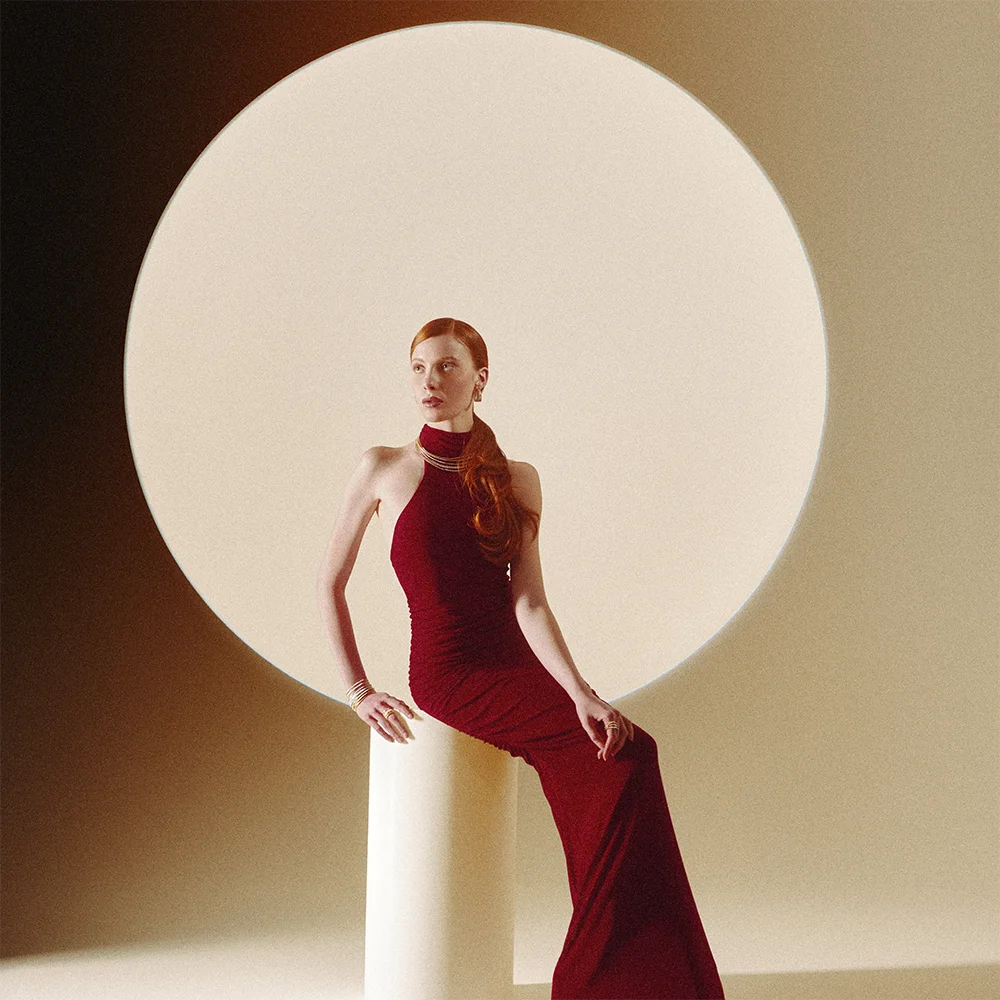 Fashion jewelry editorial 999 – model in a red halter gown wearing a gold multi-strand necklace, bracelets and rings against a circular luminous backdrop – Sbranzo Alessandro Valori Studio Vago Nicole Ciabatti