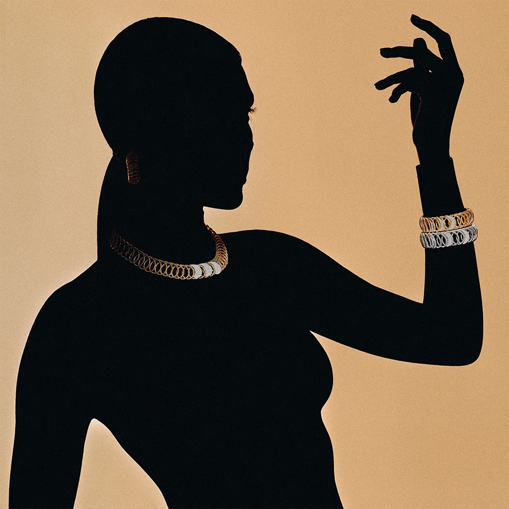 Fashion jewelry editorial 999 – silhouette of a model wearing a gold and silver chain necklace, hoop earring and stacked bracelets against a camel background – Sbranzo Alessandro Valori Studio Vago Viola Sgriscia