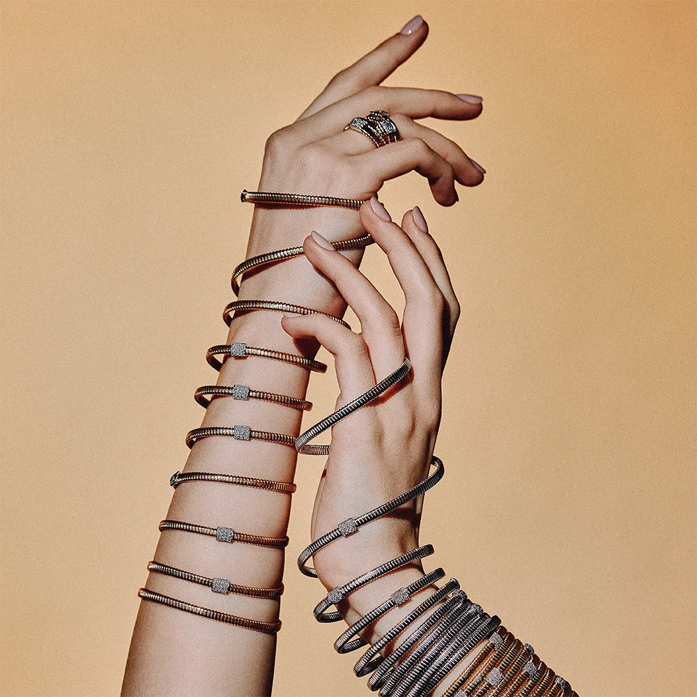 Fashion jewelry editorial – two arms covered in stacked spiral bracelets with diamond accents and a ring against a camel background – Sbranzo Alessandro Valori Studio Vago Viola Sgriscia