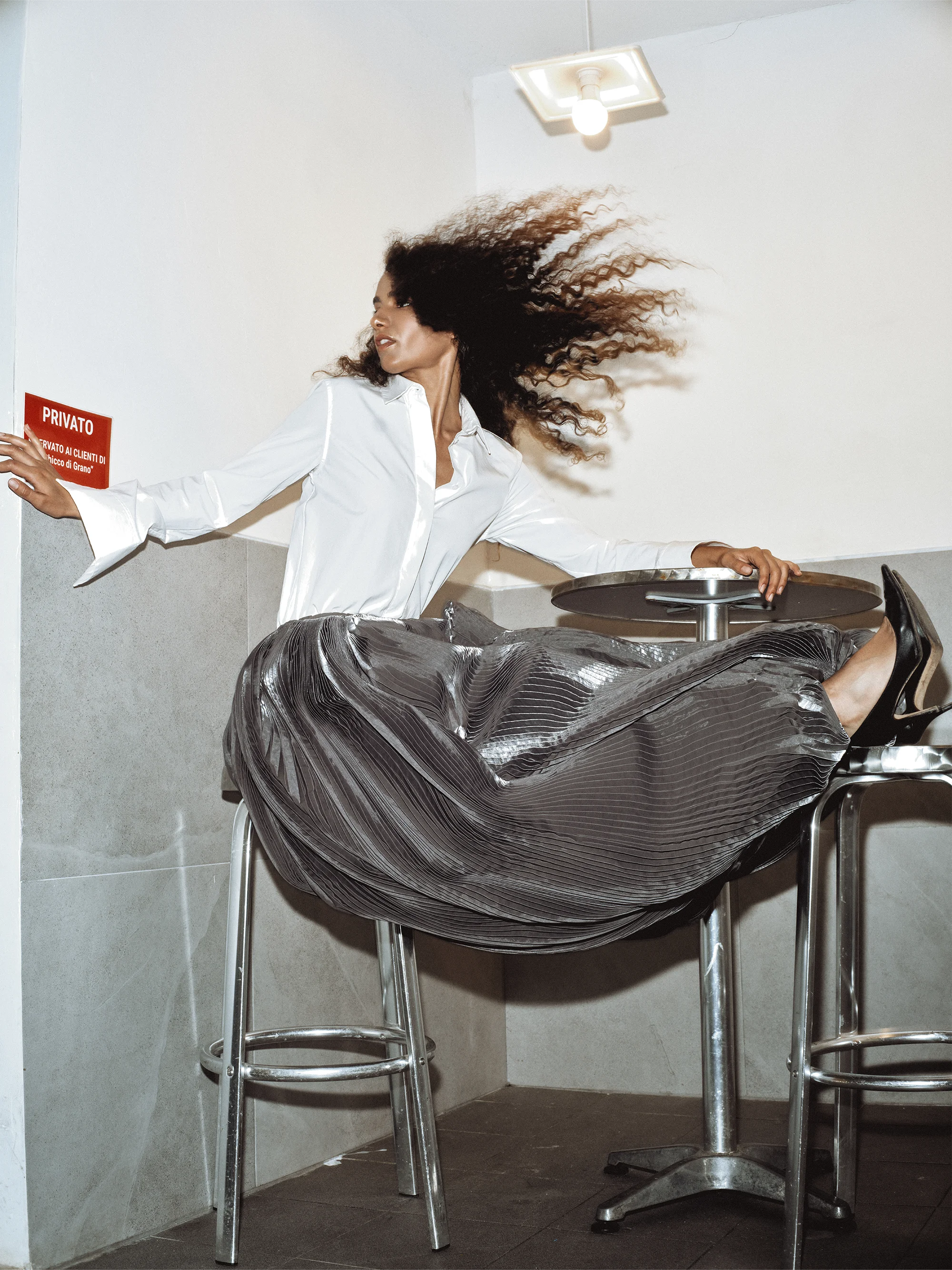 Editorial portrait of Dalila Orlandi leaning between a wall and a bar table with hair caught in motion blur, wearing a white shirt and pleated metallic skirt – Sbranzo Alessandro Valori Studio Vago Dalila Orlandi – art director Celina Ludes