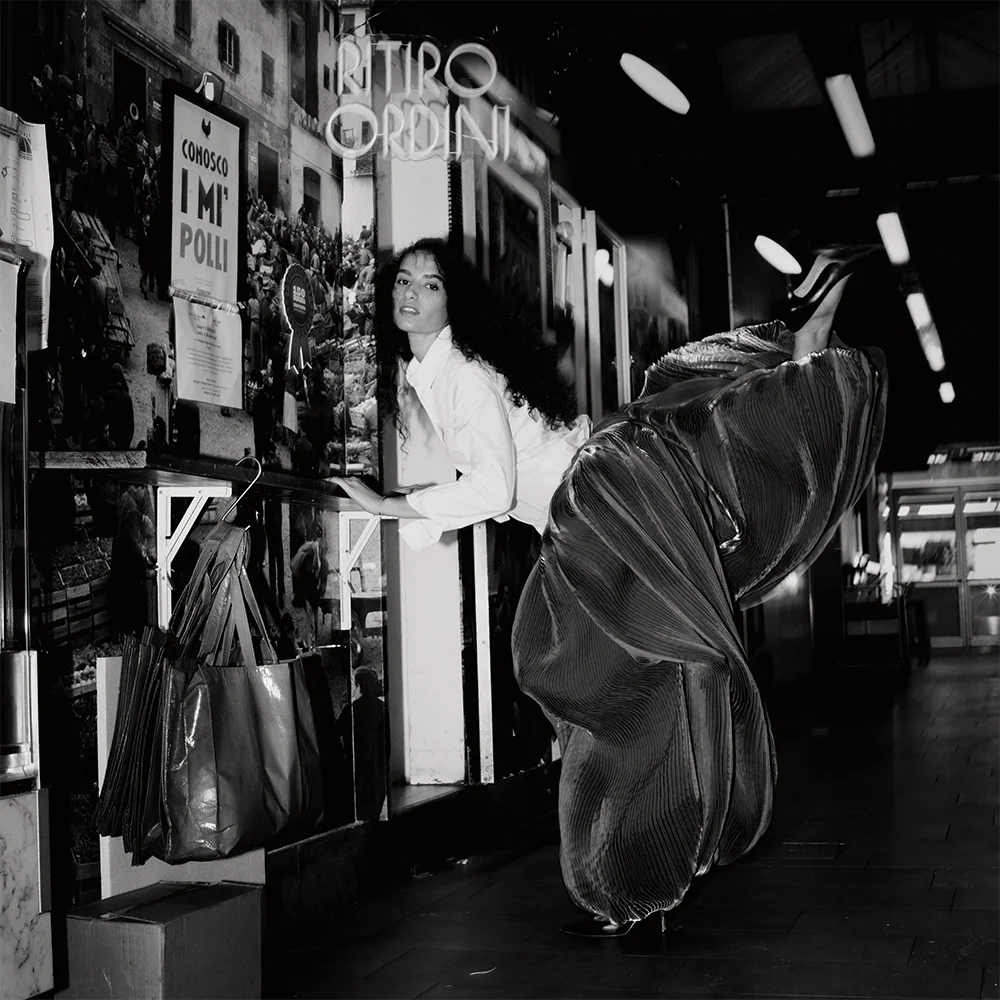 Editorial portrait of Dalila Orlandi in a suspended horizontal pose wearing a white shirt and pleated metallic skirt inside a covered market with neon signage – Sbranzo Alessandro Valori Studio Vago Dalila Orlandi – art director Celina Ludes