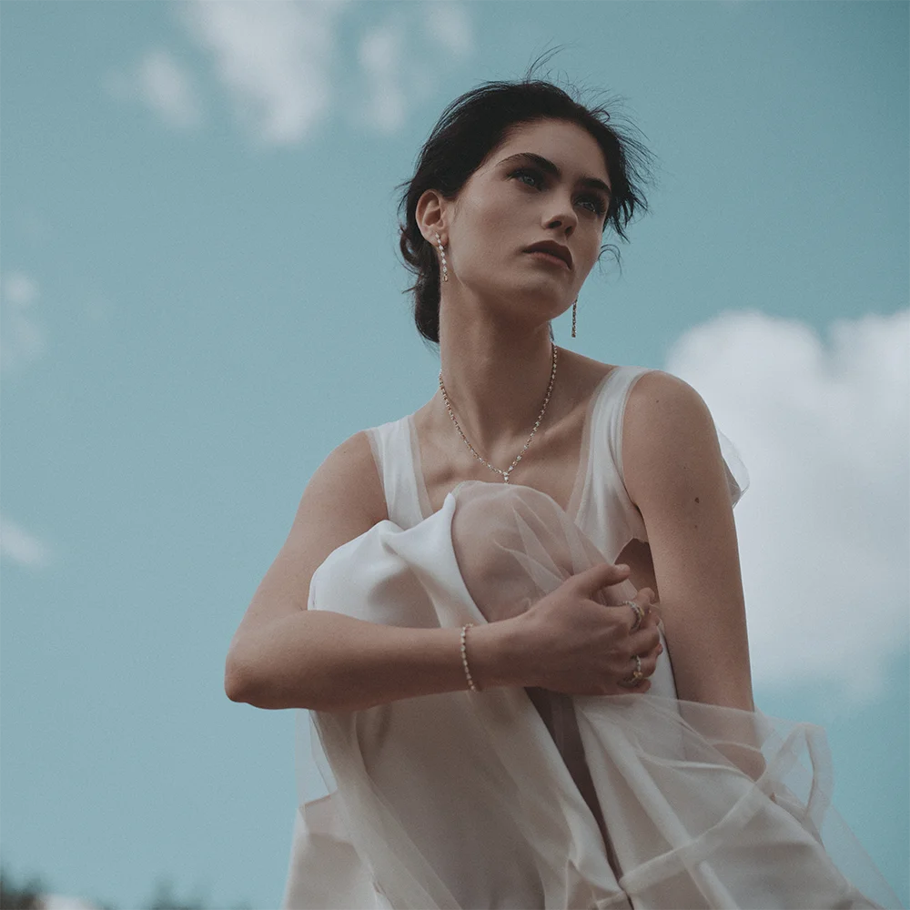 Editorial fashion jewelry portrait of a model in a sheer white dress against a blue sky wearing a diamond necklace, tennis bracelet, rings and a pendant earring – Fabiani Gioiellerie – Sbranzo Alessandro Valori Studio Vago Gaia Schiralli