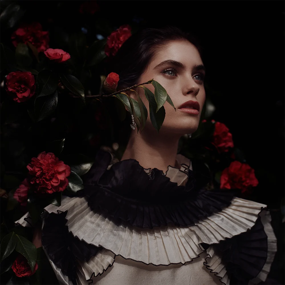 Editorial fashion jewelry portrait of a model among red camellias with a leaf branch across her face, wearing a pleated ruffle collar and drop earring – Fabiani Gioiellerie – Sbranzo Alessandro Valori Studio Vago Gaia Schiralli