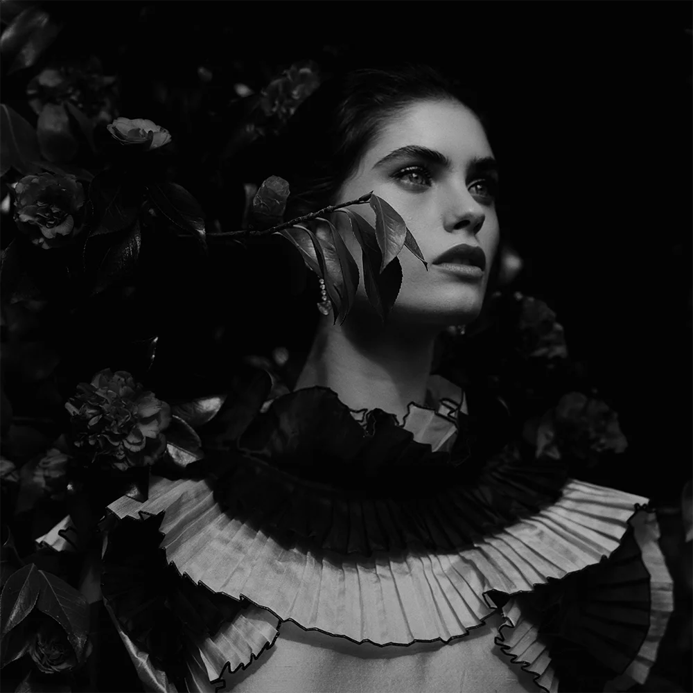 Black and white editorial fashion jewelry portrait of a model among camellias with a leaf branch across her face, wearing a pleated ruffle collar and drop earring – Fabiani Gioiellerie – Sbranzo Alessandro Valori Studio Vago Gaia Schiralli