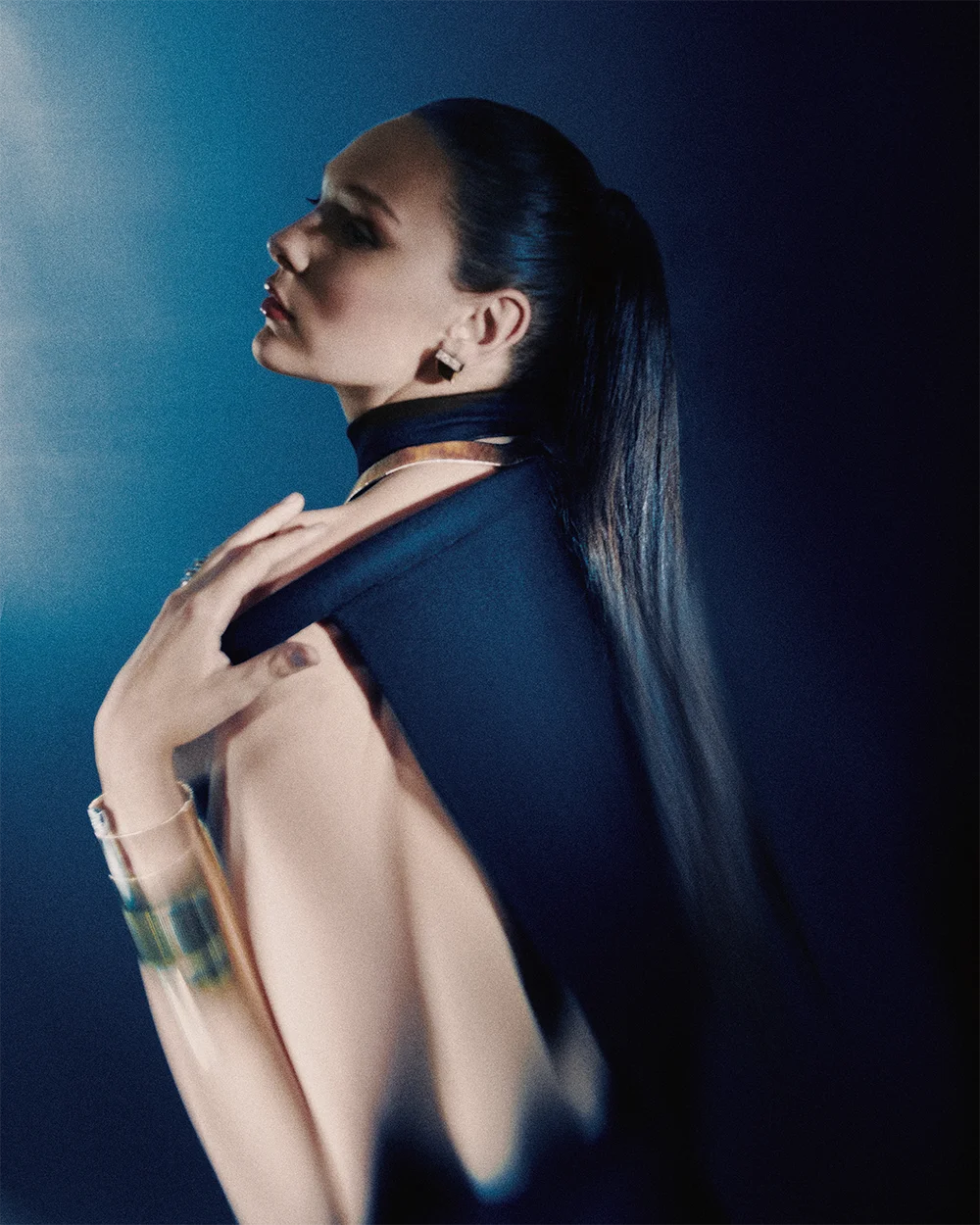 High jewelry editorial 999 – model seen from behind in profile with a long ponytail and navy cape wearing a gemstone earring and cuff bracelet against a deep blue background – Sbranzo Alessandro Valori Studio Vago Viola Sgriscia