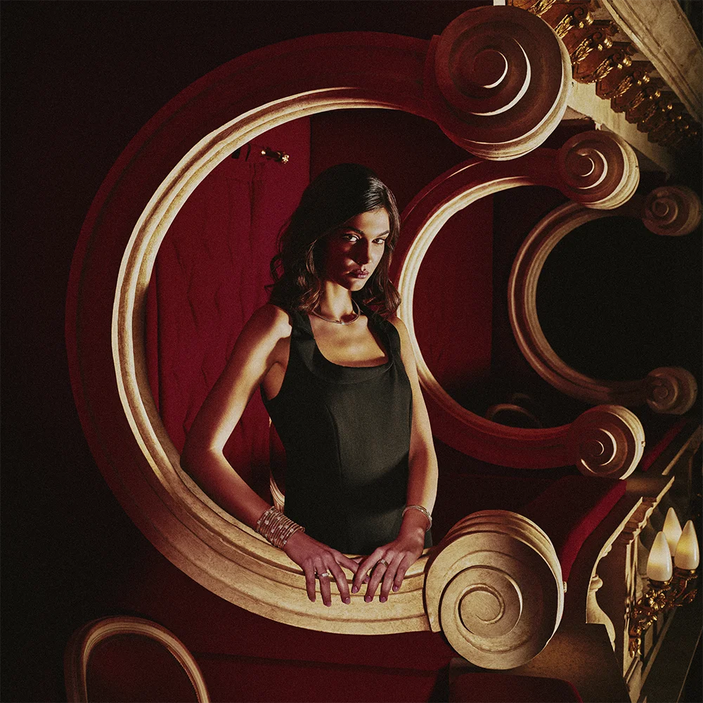 Fashion jewelry editorial 999 – model in a black dress wearing bracelets and a ring framed by an ornamental C-shaped arch in a red interior – Sbranzo Alessandro Valori Studio Vago Tati Janusheva