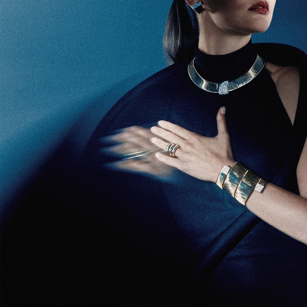 High jewelry editorial 999 – model in a navy cape dress with hand on chest wearing a gold collar necklace, gemstone earrings, a wide cuff and ring against a deep blue background crop – Sbranzo Alessandro Valori Studio Vago Viola Sgriscia