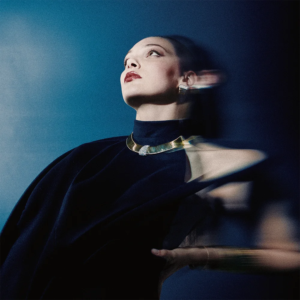 High jewelry editorial 999 – model gazing upward in a navy cape wearing a gold collar necklace with a gemstone detail and earring with a motion-blur effect against a deep blue background – Sbranzo Alessandro Valori Studio Vago Viola Sgriscia