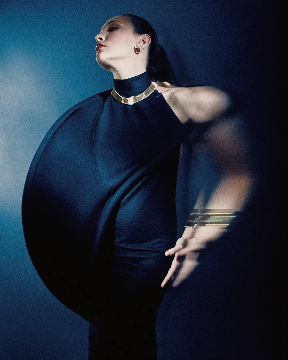 High jewelry editorial 999 – model in a navy cape gown with an outstretched motion-blurred arm wearing a gold collar necklace, earring and gold bracelets against a deep blue background – Sbranzo Alessandro Valori Studio Vago Viola Sgriscia