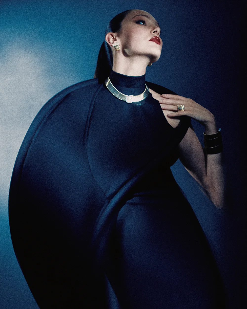 High jewelry editorial 999 – model in a sculptural navy cape gown wearing a gold collar necklace, ring and black rigid cuff against a deep blue background – Sbranzo Alessandro Valori Studio Vago Viola Sgriscia