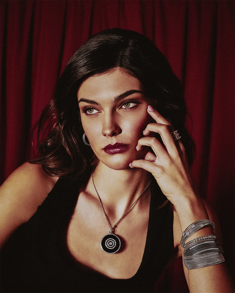 Fashion jewelry editorial 999 – close-up portrait of a model wearing a circular pendant necklace, stacked bracelets and ring against red curtains – Sbranzo Alessandro Valori Studio Vago Tati Janusheva