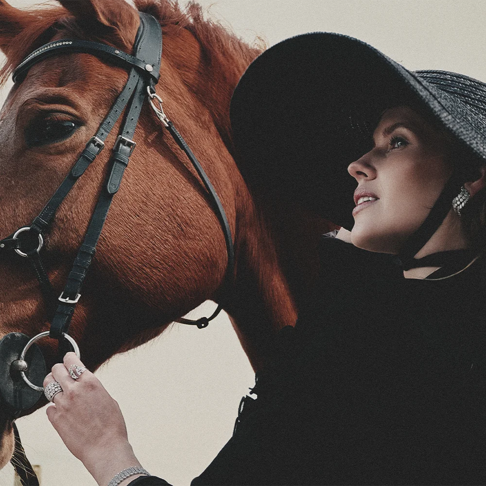 Fashion jewelry editorial – model in profile wearing a wide-brim hat and diamond rings resting her hand on a chestnut horse's bridle, with a diamond earring – Sbranzo Alessandro Valori Studio Vago